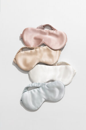 A collection of luxury silk eye masks in soft pink, champagne, ivory, and baby blue colors stacked in an elegant display on a bright background.