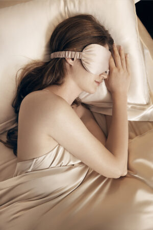 A woman lying on a silk pillow, wearing a luxurious champagne silk eye mask, epitomizing relaxation and comfort.