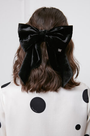 Stylish woman wearing a Duchess Silk-Velvet Bow Barrette on her hair, detailed with a monogrammed 'M', set against a polka dot sweater.